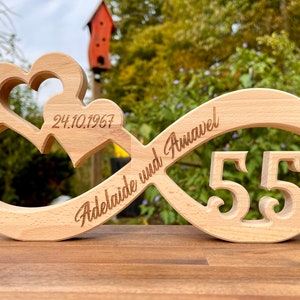 Könnte beinhalten: Ein Holz-Unendlichkeitszeichen mit zwei ineinander verschlungenen Herzen, graviert mit dem Datum "24.10.1967", den Namen "Adelaide und Annavel" und der Zahl "55".