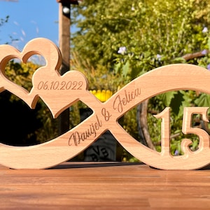 Könnte beinhalten: Holz-Unendlichkeitszeichen mit zwei Herzen, die in die Oberseite geschnitzt sind. Das Symbol ist mit dem Datum "06.10.2022", den Namen "Daniel & Felicia" und der Zahl "15" graviert.