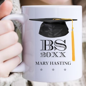 May include: White ceramic mug featuring a black graduation cap with a gold tassel. The mug has the text "BS 20XX" and the name "MARY HASTING" printed in black. Three gray stars are below the name. The mug is held by a person.