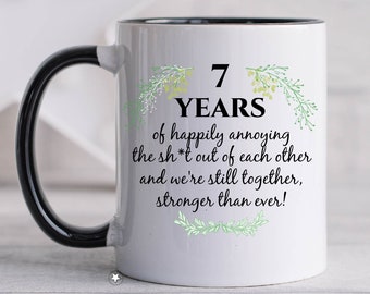 May include: White ceramic coffee mug with a black handle. The mug has a black rim and features a green floral design with the text "7 YEARS of happily annoying the sh*t out of each other and we're still together, stronger than ever!"