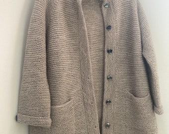 May include: A beige, knitted cardigan with a high collar and long sleeves. The cardigan has a button-up front, two front pockets, and is hanging on a hanger. The knitwear appears to be a chunky knit, with a textured surface.