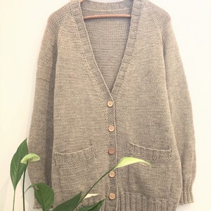 May include: A beige knitted cardigan with a V-neck, long sleeves, and two front pockets. The sweater has a button-up closure with wooden buttons and is hanging on a wooden hanger.
