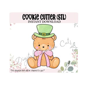 Puede incluir: Un diseño de cortador de galletas de oso de peluche de dibujos animados. El oso es de color marrón claro con un sombrero de duende verde, un lazo rosa a cuadros y una expresión linda. El texto en la parte superior dice "COOKIE CUTTER (STL) INSTANT DOWNLOAD".