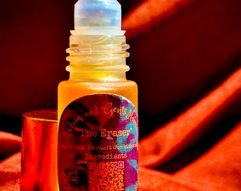 May include: A clear glass roller bottle with a silver roller ball. The bottle is labeled "Adam and Gent's Naturals The Eraser" and has a QR code. The bottle is sitting on a red fabric surface.