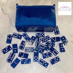 May include: A set of blue dominoes with white dots scattered around a matching blue box. The box has a lid and is open. The dominoes are made of a shiny material. The image also includes the text "Tweets Gifts & More LLC Customized Unique Gifts."