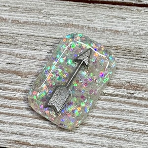 May include: A rectangular, clear resin pendant with iridescent glitter and a silver arrow design. The arrow points upwards, set against a backdrop of colorful, sparkling glitter. The pendant is displayed on a weathered, light-colored wooden surface.