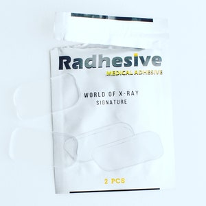 May include: A white packet of Radhesive medical adhesive with the text "WORLD OF X-RAY SIGNATURE" and "2 PCS". Two clear adhesive pads are visible, along with a partially opened top.
