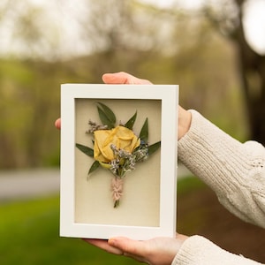 May include: A white frame with a pressed yellow rose and greenery inside. The frame is being held by a person's hand.