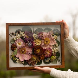 May include: A framed floral arrangement with dried flowers in pink, purple, and yellow hues. The flowers are arranged on a light fabric and enclosed in a wooden frame. The artwork is held up against a blurred green background.