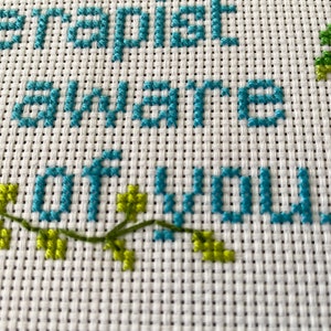 May include: Cross-stitch embroidery with the text "The happiest is aware of you" in teal blue thread. Green leaves are stitched on the bottom left corner and a red, green, and yellow heart is stitched on the top right corner.