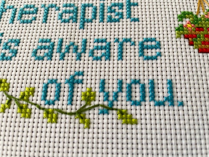 May include: Cross-stitch embroidery with the text "The happiest is aware of you" in teal blue thread. Green leaves are stitched on the bottom left corner and a red, green, and yellow heart is stitched on the top right corner.