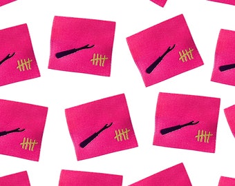 May include: Pink fabric squares with black and gold embroidery. The embroidery depicts a small black tool with a handle and a small gold fence with three vertical lines. The text "HHH" is embroidered in gold below the fence.