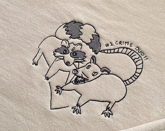 Peut inclure: Un sweat-shirt beige avec un motif brodé de deux ratons laveurs avec le texte "#1 CRIME DUO" au-dessus.