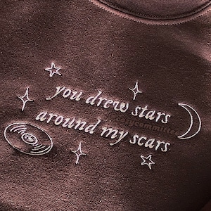 May include: A brown sweatshirt with white embroidery that reads "you drew stars around my scars". The embroidery also includes a crescent moon and several stars.