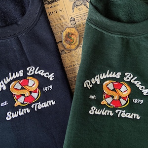 以下が含まれることがあります： 「Regulus Black est. 1979 Swim Team」と白く刺繍された黒いスウェットシャツと緑のスウェットシャツ2枚。デザインは、白とオレンジのライフセーバーの中に蛇が入っている。