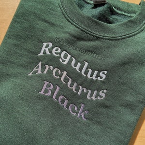 以下が含まれることがあります： 暗い緑色のスウェットシャツに、薄いグレーの刺繍で「Regulus Arcturus Black」という文字が書かれています。