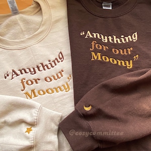 Op de afbeelding: Twee sweatshirts met de tekst "Anything for our Moony" geborduurd in goud op een bruine en beige achtergrond. Het bruine sweatshirt heeft een halve maan geborduurd op de mouw en het beige sweatshirt heeft een ster geborduurd op de mouw. De tekst "@cozycommittee" is zichtbaar op het bruine sweatshirt.