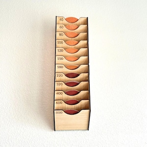 May include: A wooden sandpaper organizer with multiple compartments, each labeled with grit numbers from 40 to 800. The compartments hold sandpaper in shades of orange and red. The organizer is made of light-colored wood.