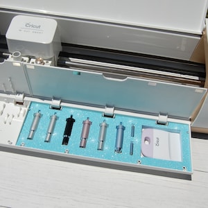 May include: Open Cricut Smart cutting machine with a blue glitter mat and a variety of different colored blades and tools. The blades are silver, black, pink, and blue. The tools include a scraper and a small metal tool.