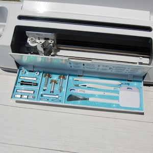 May include: An open white Cricut Explore Air 2 machine with the tool tray open. The tray is filled with various tools, including a spatula, scraper, tweezers, and a variety of blades. The tray is lined with a blue glitter material.