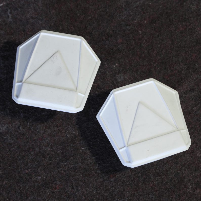 May include: Two white, geometric-shaped objects with a triangular design in the center. The objects have a multi-faceted, octagonal shape. The items are displayed on a dark brown, textured surface.