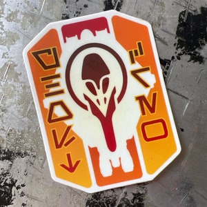 May include: Orange and red sticker with a white alien-like creature in the centre. The creature has a red head and a white body. The sticker has the text "DEVONO" on the right side.