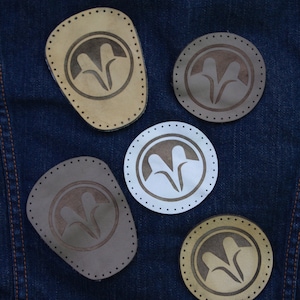 May include: Five leather patches with a stylized design of two stylized leaves. The patches are brown, beige, and white. The patches are on a blue denim background.
