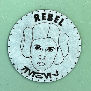 May include: A silver round leather patch with a black outline of Princess Leia's face and the words "Rebel" and "Yewj" in black.