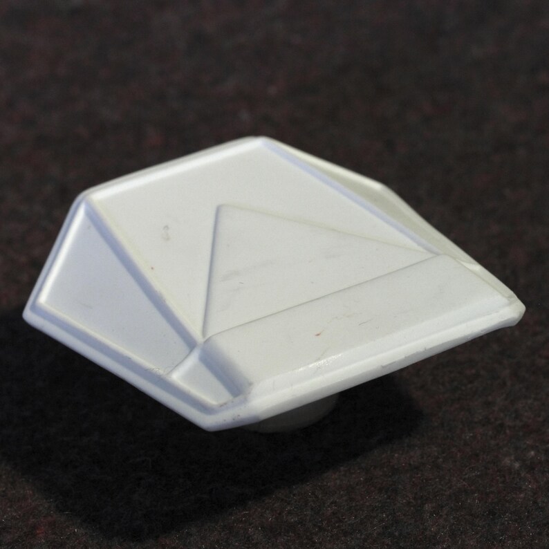 May include: A white, geometric cabinet knob with a faceted design. The knob has a square shape with beveled edges and a triangular accent. The knob is made of a solid, opaque material and has a small, round base.