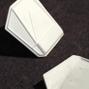 May include: Two white, geometric-shaped objects are displayed against a dark brown background. One object is a three-dimensional, angular form with a triangular design. The other object is a flat, hexagonal shape.