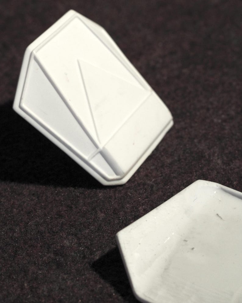 May include: Two white, geometric-shaped objects are displayed against a dark brown background. One object is a three-dimensional, angular form with a triangular design. The other object is a flat, hexagonal shape.