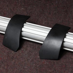 May include: Two black, curved, plastic pieces are positioned on a silver metal bar. The pieces have a matte finish and are set against a brown, textured background. The design suggests a possible automotive or industrial component.