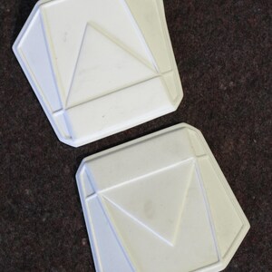 May include: Two white, geometric-shaped objects with a pentagonal outline and a central triangle design. The objects have a raised, textured surface and are set against a dark brown background.