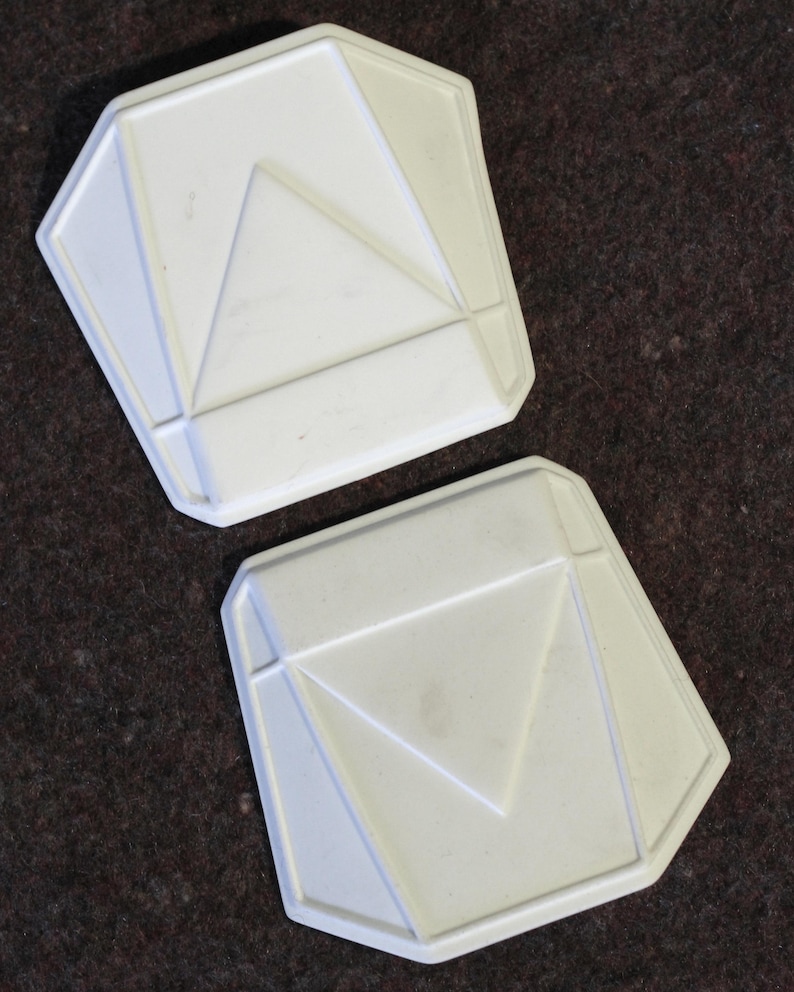 May include: Two white, geometric-shaped objects with a pentagonal outline and a central triangle design. The objects have a raised, textured surface and are set against a dark brown background.