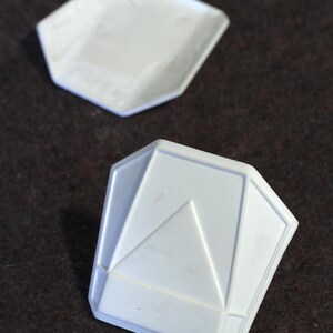 May include: Two white, geometric-shaped dishes. The top dish is a simple, shallow tray. The bottom dish has a raised, angular design with a central triangle. Both are set against a dark brown background.