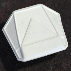 May include: A white, geometric-shaped object with a triangular design in the center. The object has a raised, beveled edge and a matte finish. The shape is a modified octagon. The object appears to be made of ceramic or plastic.