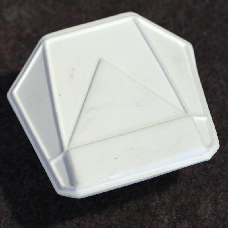 May include: A white, geometric-shaped object with a triangular design in the center. The object has a raised, beveled edge and a matte finish. The shape is a modified octagon. The object appears to be made of ceramic or plastic.