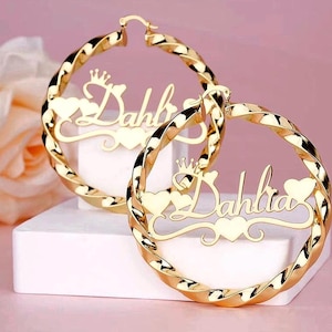 May include: A pair of gold-toned hoop earrings featuring a twisted metal design. Each earring displays the name Dahlia in a cursive script, accented with a small crown above the name and heart shapes integrated into the lettering and frame.