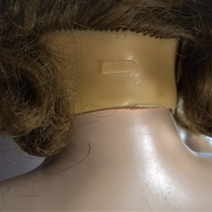 Op de afbeelding: Close-up van de achterkant van een poppenhoofd met bruin haar. De nek van de pop heeft een markering met de tekst "Dollcraft".