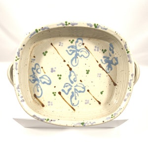 May include: A handmade, off-white ceramic baking dish with a square shape and rounded corners. It features blue floral designs, brown stems, and green and purple accents. The dish has two handles and is ideal for serving.