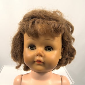 Op de afbeelding: Een vintage poppenhoofd met bruin krullend haar en blauwe ogen. De pop heeft een geschilderd gezicht met roze wangen en een gesloten mond.