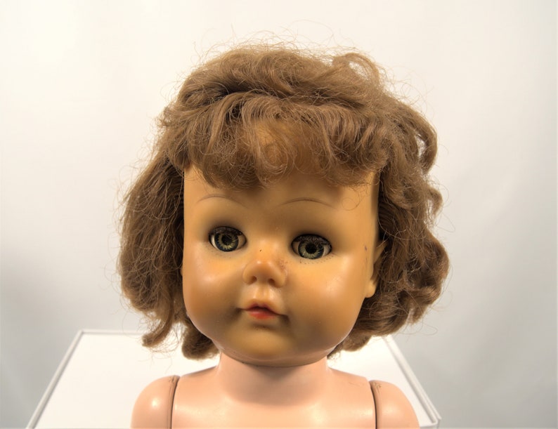 Op de afbeelding: Een vintage poppenhoofd met bruin krullend haar en blauwe ogen. De pop heeft een geschilderd gezicht met roze wangen en een gesloten mond.