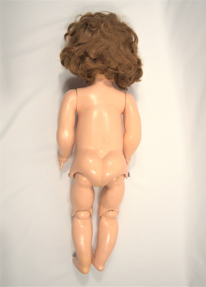 Op de afbeelding: Een vintage porseleinen pop met bruin krullend haar en een naakt lichaam. De pop heeft scharnierende armen en benen.