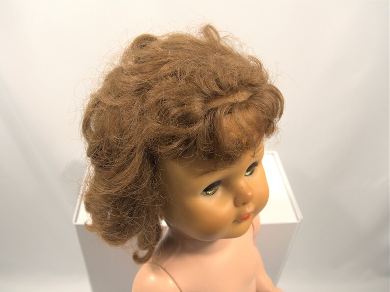 Op de afbeelding: Een vintage pop met bruin krullend haar en blauwe ogen. De pop draagt een naakt lichaam en heeft een geschilderde glimlach.