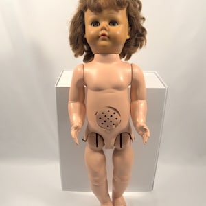 Op de afbeelding: Een vintage plastic pop met bruin krullend haar, blauwe ogen en een roze lichaam. De pop heeft een gat in zijn buik met een luidspreker.