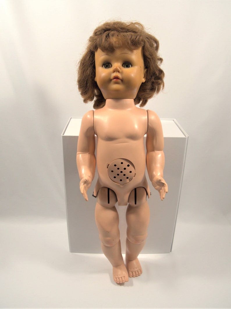Op de afbeelding: Een vintage plastic pop met bruin krullend haar, blauwe ogen en een roze lichaam. De pop heeft een gat in zijn buik met een luidspreker.