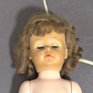 Op de afbeelding: Een vintage pop met bruin krullend haar en een geschilderd gezicht. De pop heeft gesloten ogen en een rode geschilderde mond. De pop draagt een witte jurk.