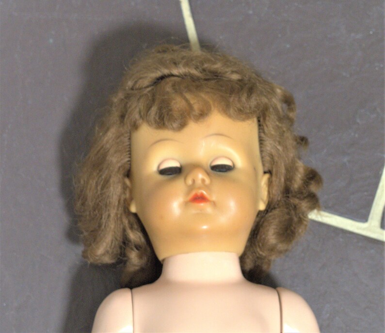Op de afbeelding: Een vintage pop met bruin krullend haar en een geschilderd gezicht. De pop heeft gesloten ogen en een rode geschilderde mond. De pop draagt een witte jurk.