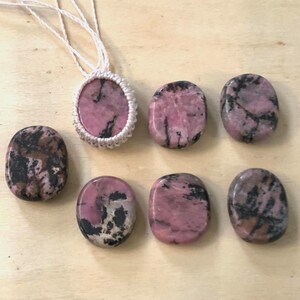 May include: Pink and black rhodonite stones in various sizes. One stone is set in a macrame pendant with a white cord. The other stones are oval-shaped and display unique patterns. The stones are displayed on a light wood surface.