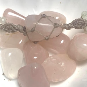May include: Close-up of a collection of polished rose quartz stones in varying shades of pink. Some stones are held in a gray macrame net. The stones are smooth and rounded, with a translucent quality. The overall aesthetic is soft and natural.